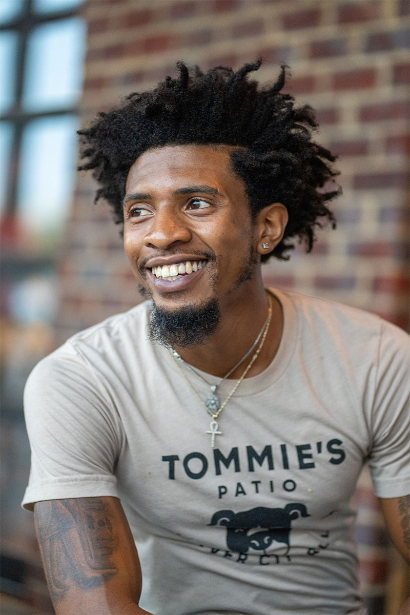 Person in t-shirt and necklaces sitting in front of brick wall.